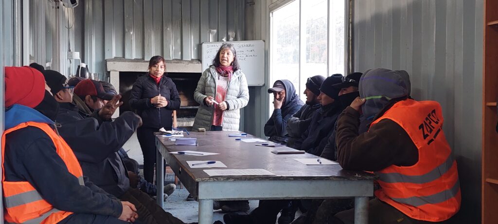 Una mujer está en una reunión presencial con los trabajadores. Ella está parada y al rededor de una mesa están sentados los hombres. Sobre la mesa hay papel y lapiceras. Detrás de la mujer hay un pizarrón que tiene escrito "Diversidad de Género"